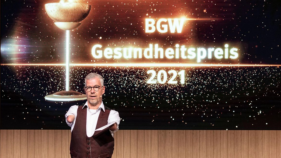  Moderator Rainer Schmidt vor einer Leinwand mit Text "BGW Gesundheitspreis 2021"