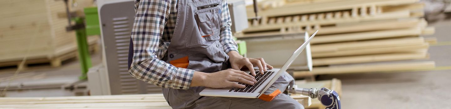 Behinderung und Arbeit Junger Arbeiter mit Beinprothese sitzt auf einem Holzstapel in einer Werkstatt und arbeitet an einem Notebook.
