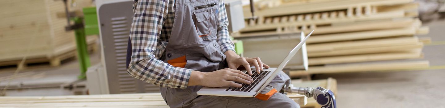 Junger Arbeiter mit Beinprothese sitzt auf einem Holzstapel in einer Werkstatt und arbeitet an einem Notebook.