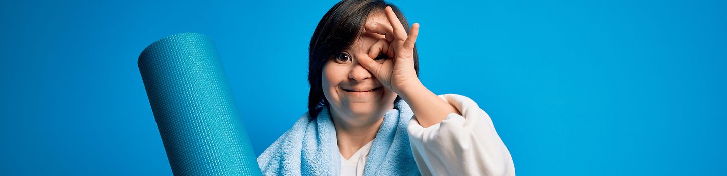 Junge Frau mit Trisomie 21 trägt eine Yogamatte und ein Handtuch. Sie lächelt, zeigt ein ok-Zeichen mit der Hand und schaut dabei durch ihre Finger.