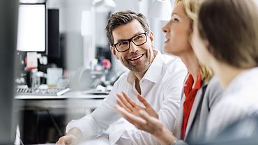 Drei Personen sitzen in einem Büroraum nebeneinander an einem Tisch