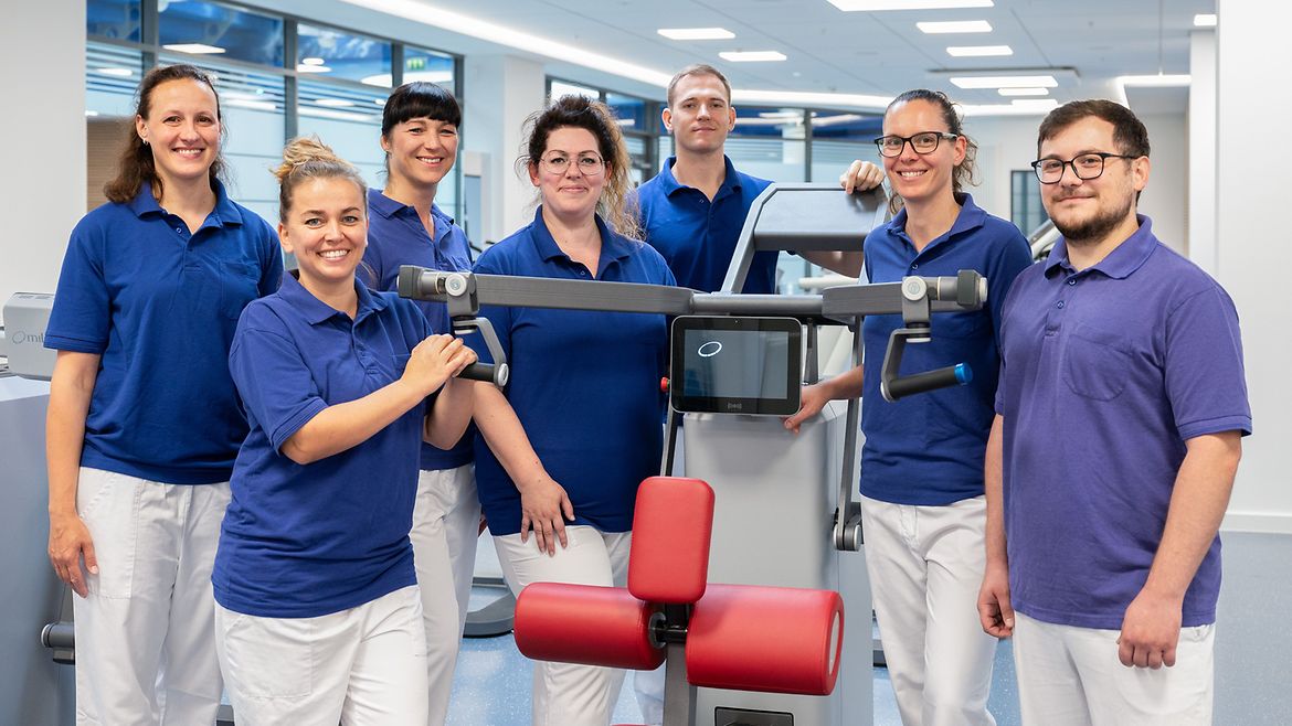 Eine Menschengruppe, die alle blaue Oberteile und weiße Hosen tragen, stehen rechts und links sowie hinter einem rot gepolsterten Fitnessgerät.