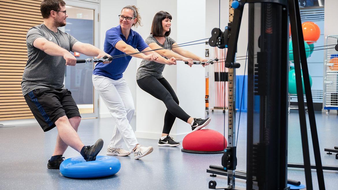 Ein Mann und eine Frau üben jeweils an einer Seilzugmaschine im Fitnessraum. Eine Therapeutin steht links neben dem Mann und korrigiert seine Haltung.