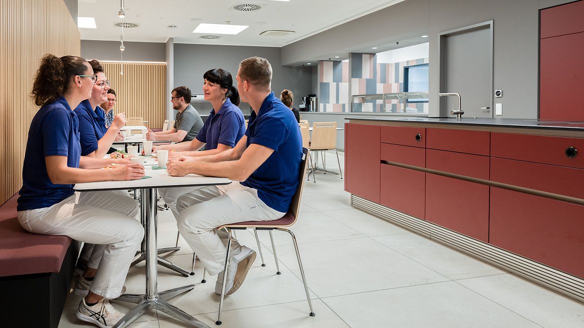 An einem Esstisch in einer Kantine unterhalten sich vier Personen. Sie tragen alle weiße Hosen und blaue Oberteile. Rechts steht eine dunkelrote Küchenzeile mit Küchentresen.