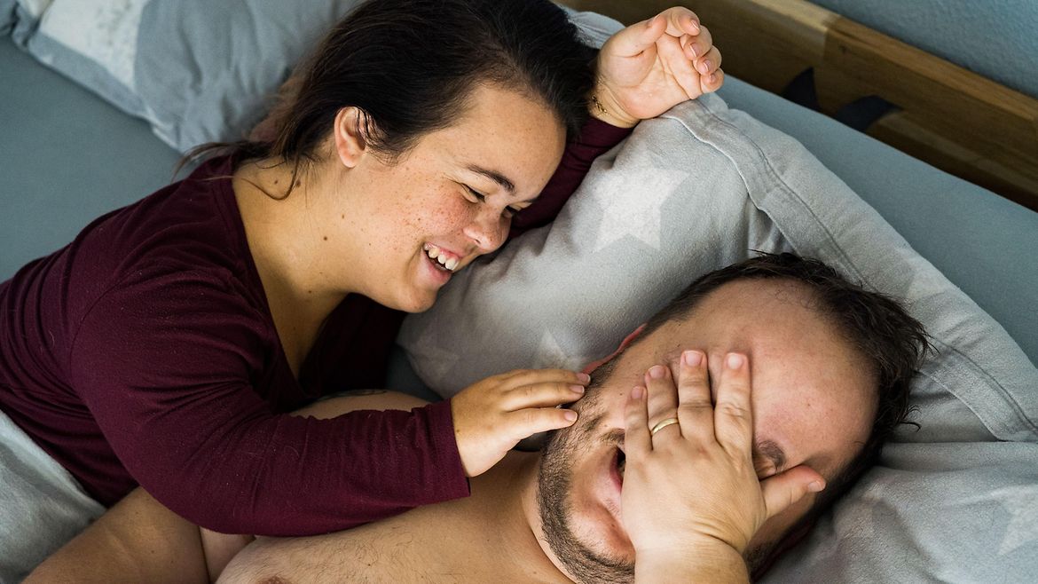 Ein junges Paar liegt nebeneinander im Bett. Sie streicht im zärtlich über die Wange. Er hat eine Hand vor dem Gesicht. Beide lachen.