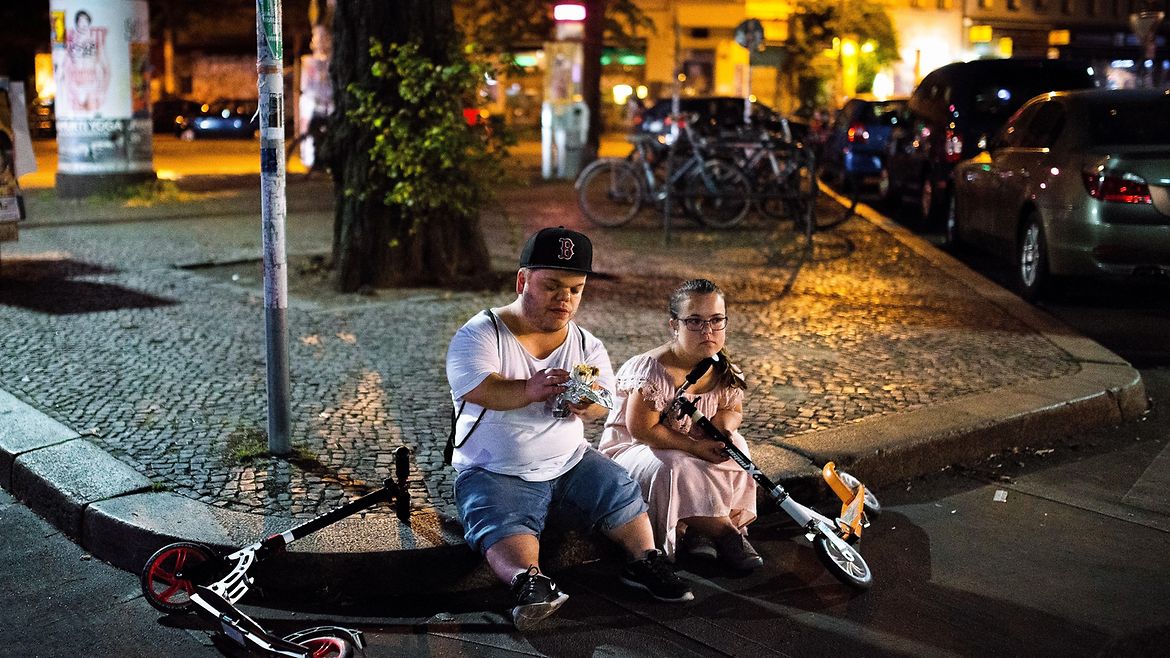 Ein kleinwüchsiges Paar sitzt auf der Bordsteinkante einer Stadt. Es ist dunkel. Neben ihnen liegen zwei Roller.
