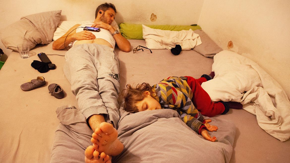 Auf einem großen Bett liegen ein Mann und ein Kind - beide schlafen. Drum herum liegen viele Gegenstände wie Schuhe, Brille, Mobiltelefon und ein Wasserglas.
