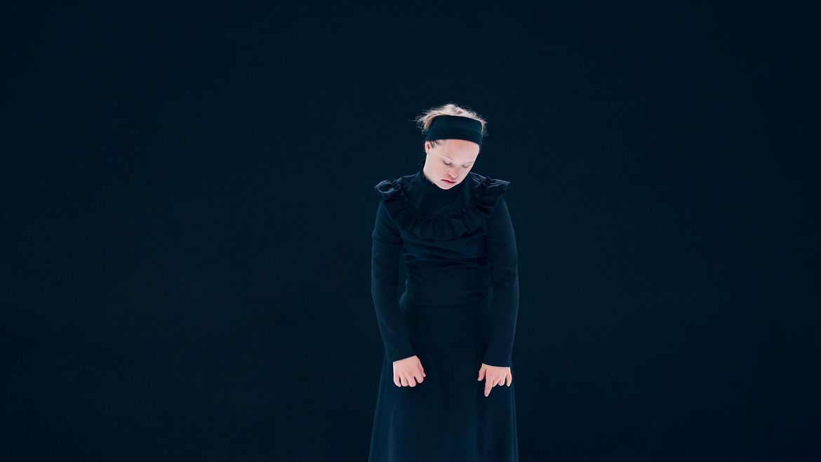 Eine Frau trägt eine schwarzes Kleid vor schwarzem Hintergrund. Man sieht nur ihr Gesicht, ihre Haare und Hände deutlich.
