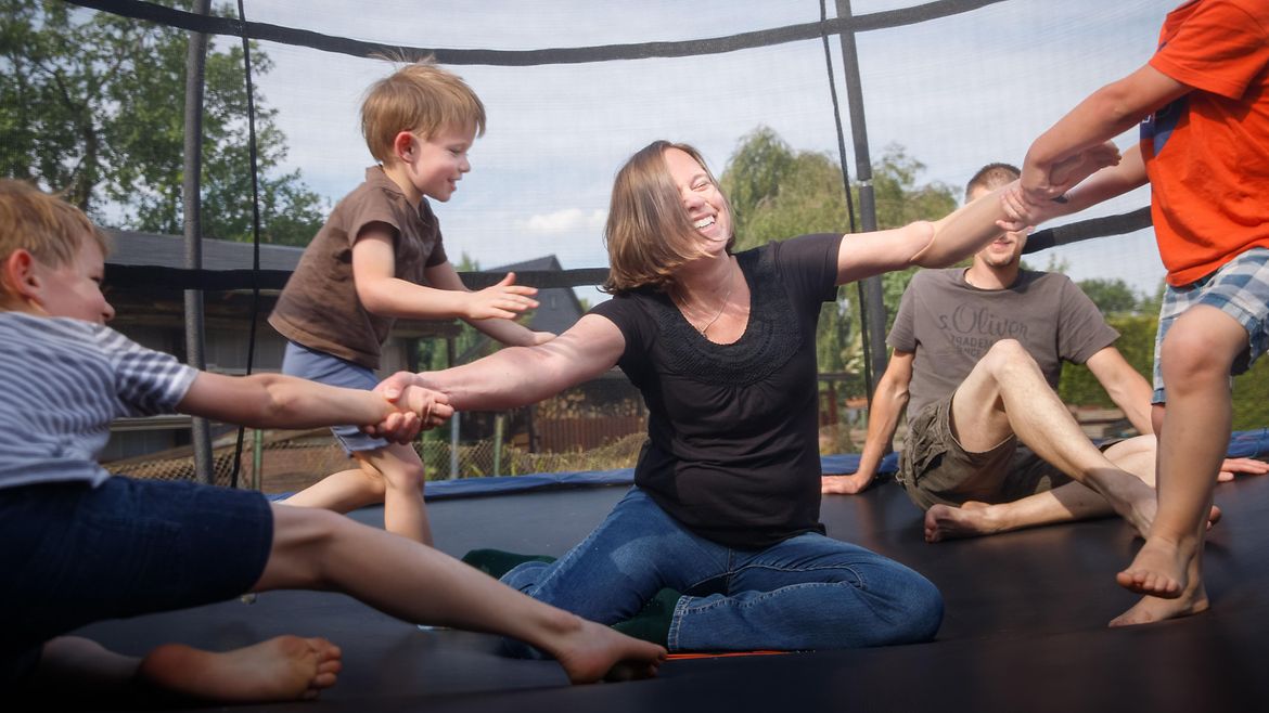 Eine Frau sitzt auf einem großen Trampolin. Um sie herum springen drei Kinder. Hinter ihr sitzt ein Mann.