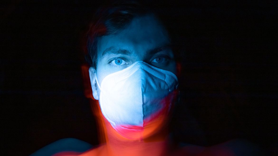 Ein Gesicht mit Mund-Nasen-Maske im Licht vor schwarzem Hintergrund.