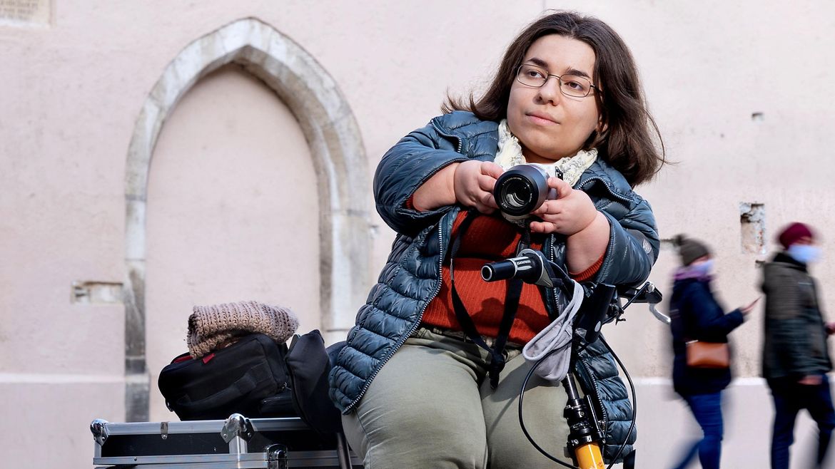 Eine kleinwüchsige Frau auf einem Roller steht auf einem Platz in der Stadt. Sie hält eine Kamera in der Hand.