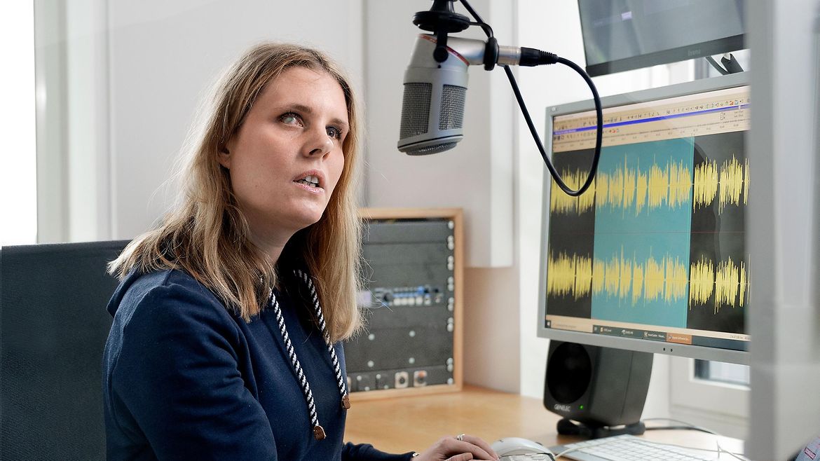 Eine blinde junge Frau sitzt in einem Radiostudio und spricht in ein Mikrofon. Sie hat die Hände auf einem Gerät abgelegt. 