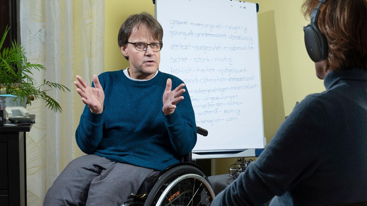 Ein Mann im Rollstuhl sitzt vor einem Flipchart und spricht mit einer weiteren Person, die Kopfhörer trägt.