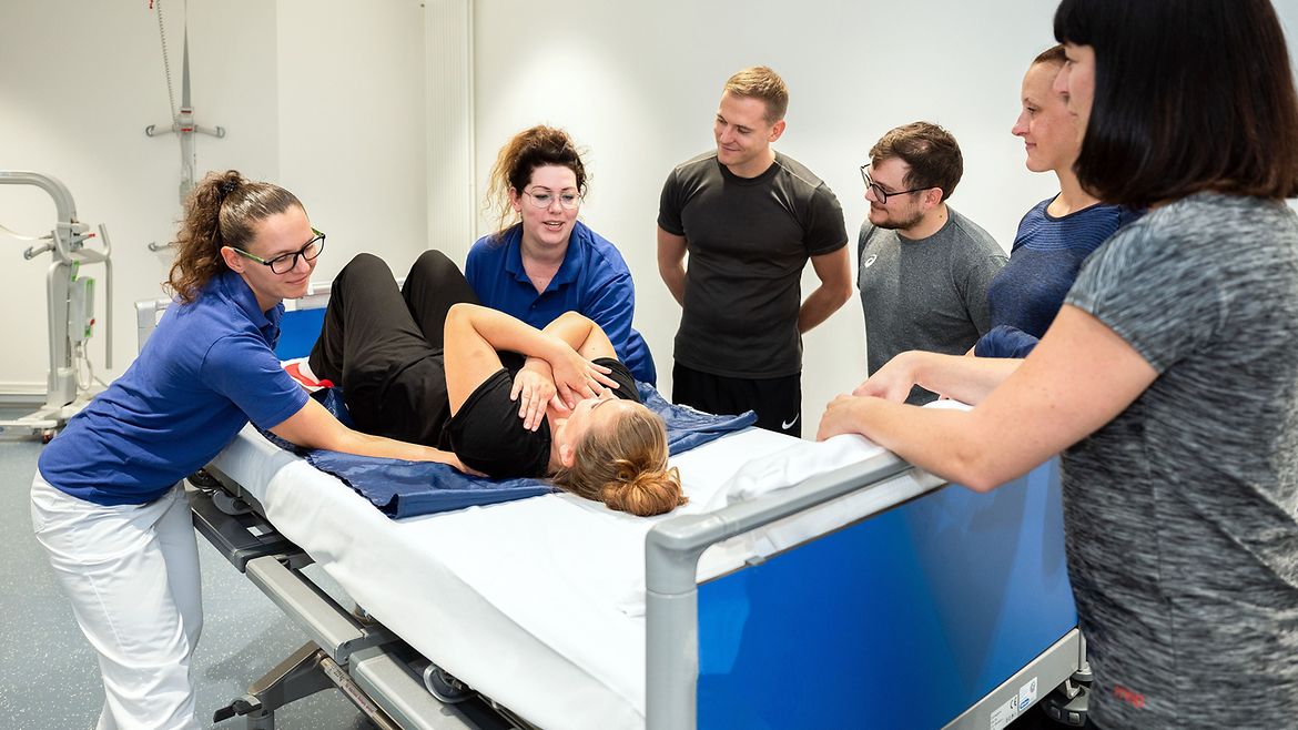Zwei Therapeutinnen zeigen, wie eine Patientin, die auf dem Rücken im Pflegebett liegt, unter Einsatz einer Rutschmatte vom unteren Bettende nach oben transferiert wird.