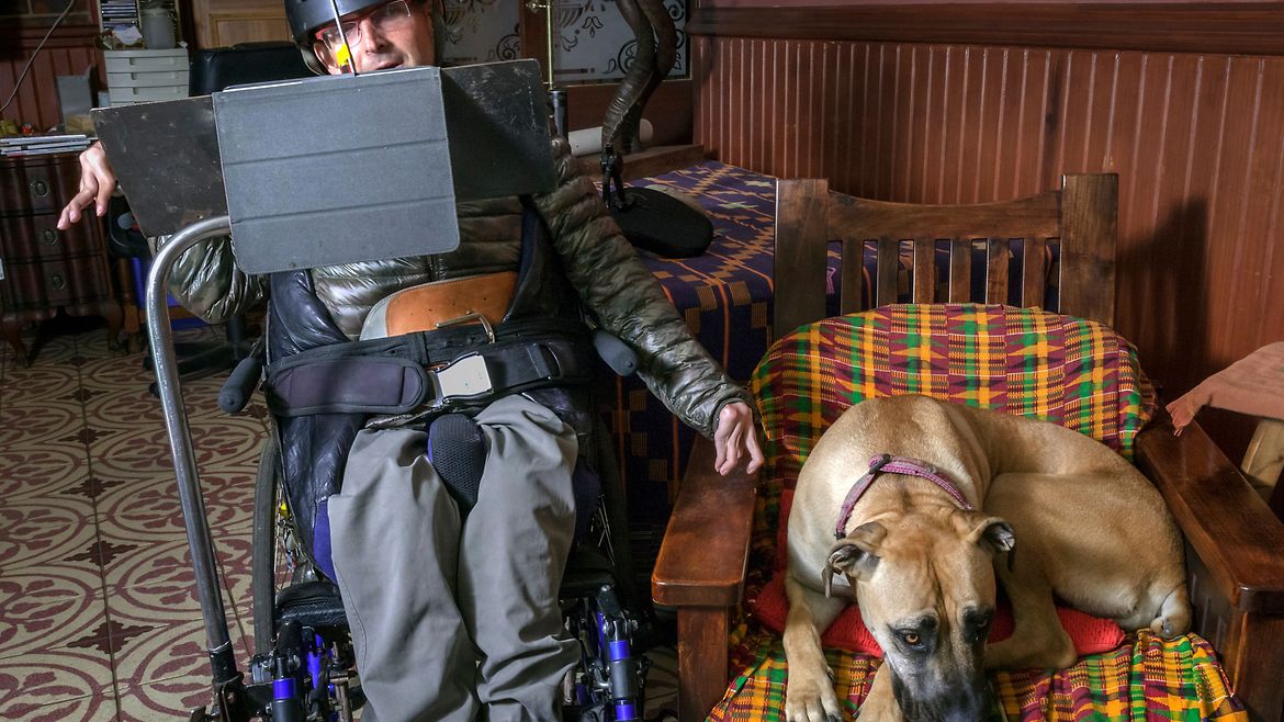 Ein spastisch gelähmter Mann sitzt im Rollstuhl. Auf dem Kopf hat er einen Helm und vor sich einen Bildschirm. Daneben im Sessel liegt ein großer brauner Hund.