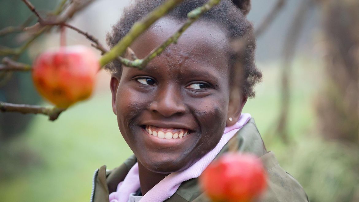 Junge Frau mit dunkler Hautfarbe auf eine Wiese. Vor hier hängen rote Äpfel an einem Baum. Sie lächelt.