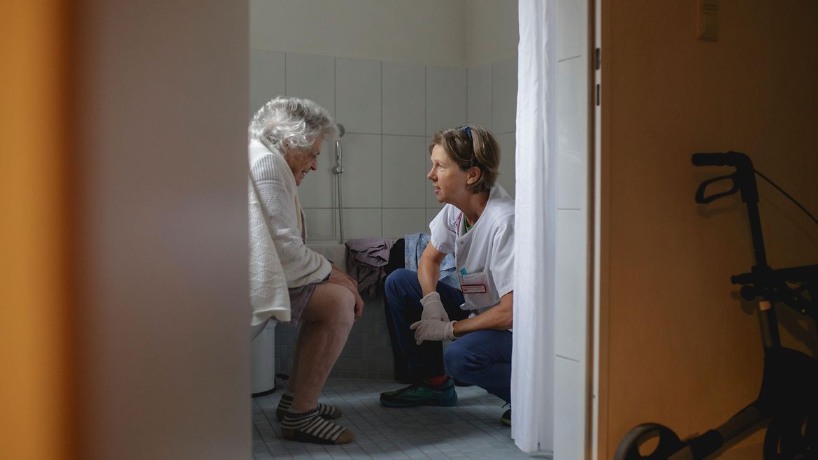 Eine Patientin sitzt auf der Toilette. Eine Pflegerin kniet ihr gegenüber.