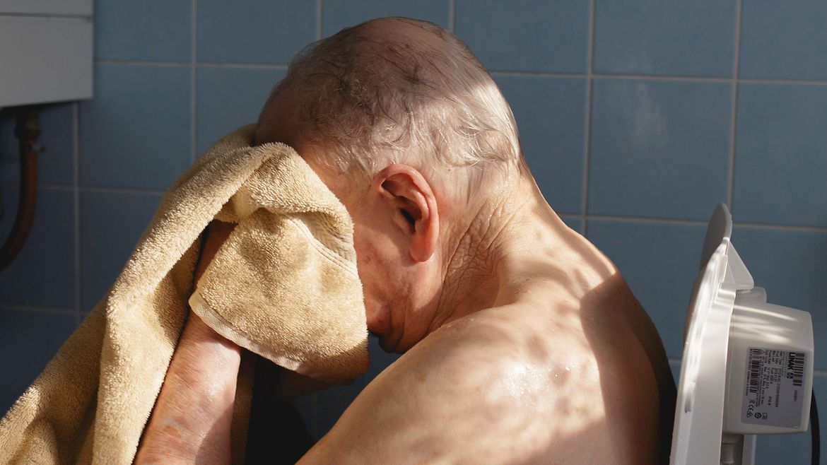 Ein älterer Mann sitzt mit nacktem Oberkörper in einem gekachelten Raum. Er trocknet sich mit einem Handtuch das Gesicht.