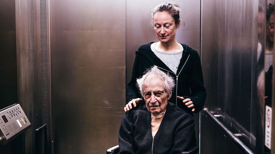 Eine Frau und ein älterer Mann im Rollstuhl befinden sich in einem Fahrstuhl.