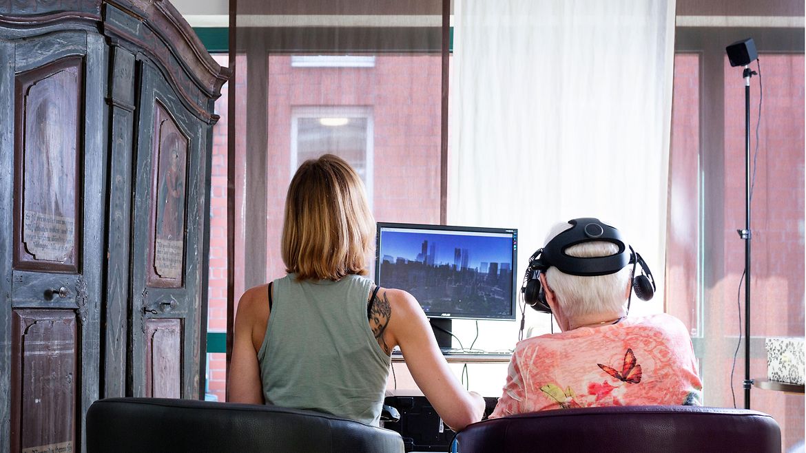 Eine ältere Frau mit VR-Brille sitzt neben einer jungen Frau vor einem Computer.