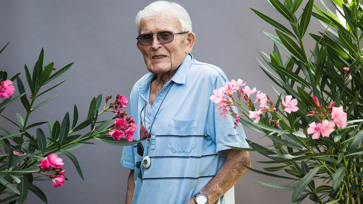 Ein älterer Mann steht mit Stock zwischen Pflanzen vor einer grauen Wand.