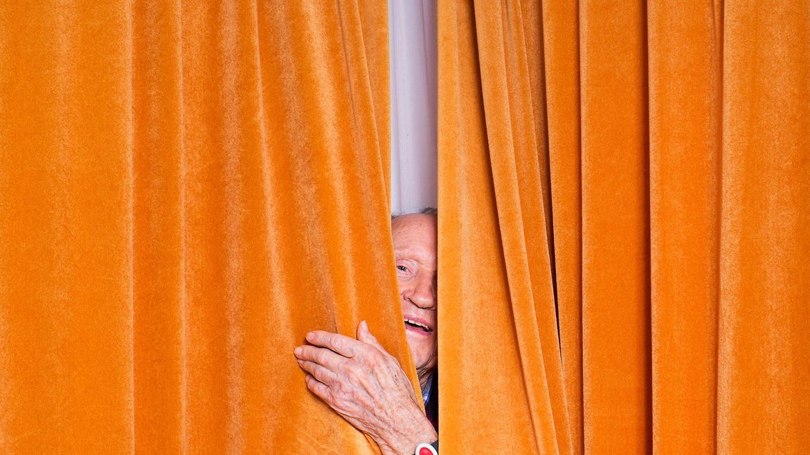 Ein älterer Mann schaut durch einen Schlitz in einem orangenen Vorhang.