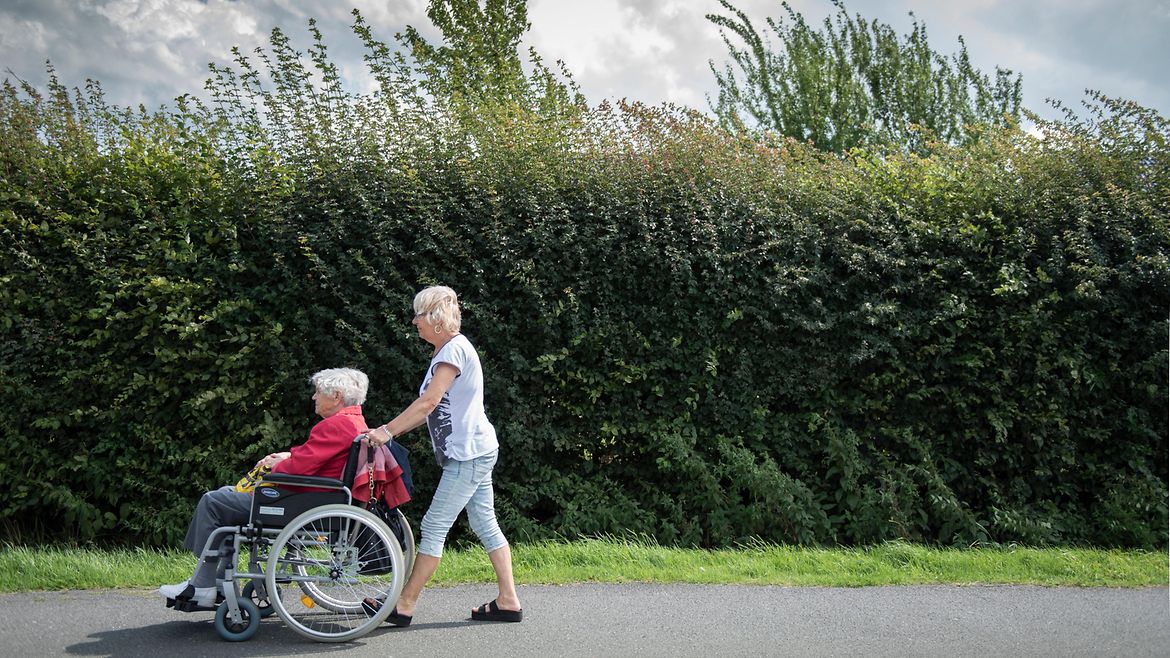 Eine junge Frau schiebt eine alte Dame im Rollstuhl an einer Hecke entlang. Im Hintergrund ziehen Wolken auf.