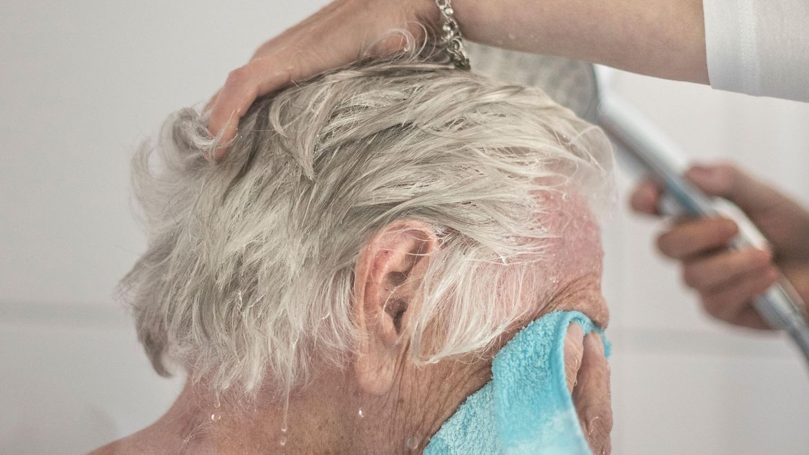 Ein älterer Mann hält sich ein Handtuch vor das Gesicht, während seine Haare gewaschen werden.