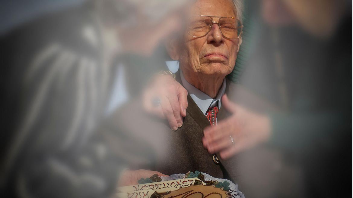 Ein älterer Herr mit Hemd und Krawatte bekommt eine Torte überreicht - verziert mit der Zahl 100.