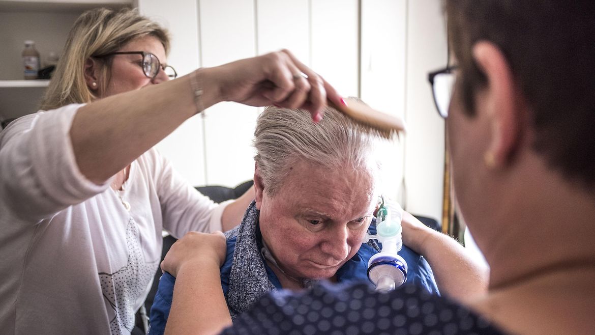 Eine Frau steht hinter einem älteren Mann und kämmt seine Haare. Eine andere Frau steht vor ihm und hält ihn an den Schultern.