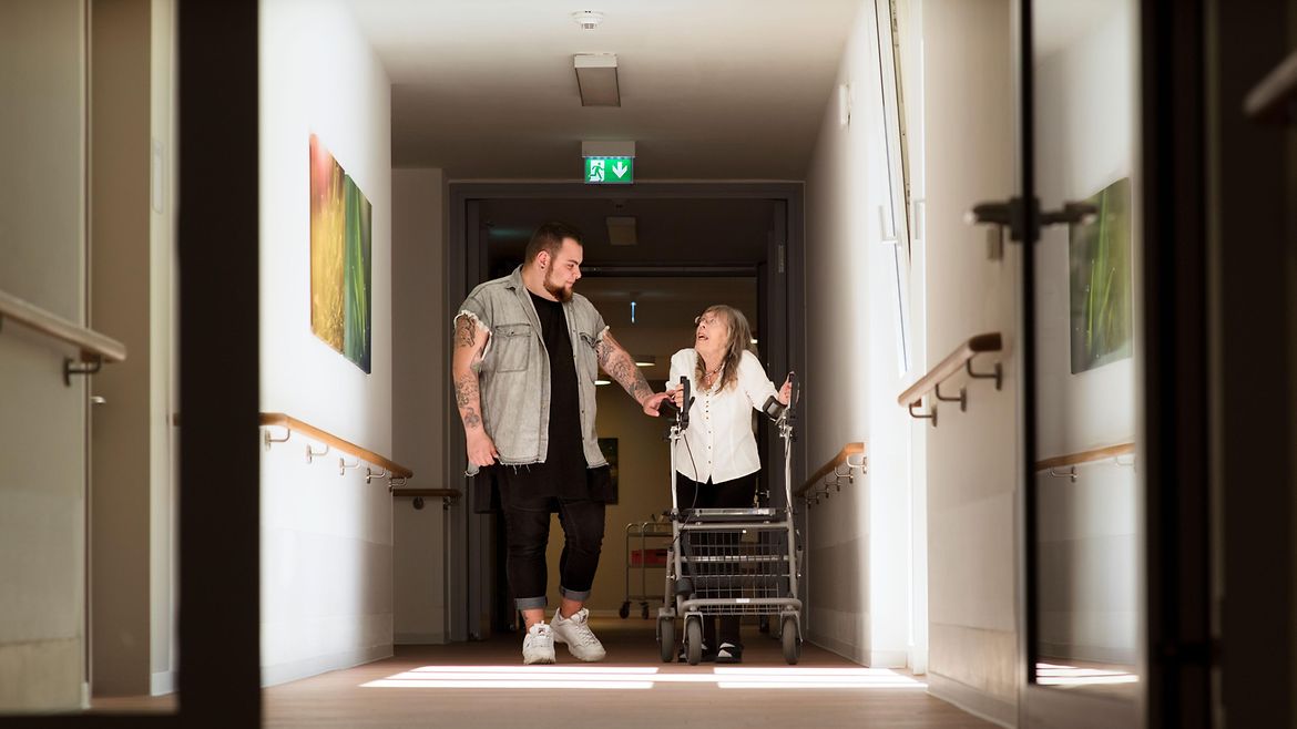 Ein Pfleger geht mit einer älteren Frau mit einem Rollator durch einen Flur.