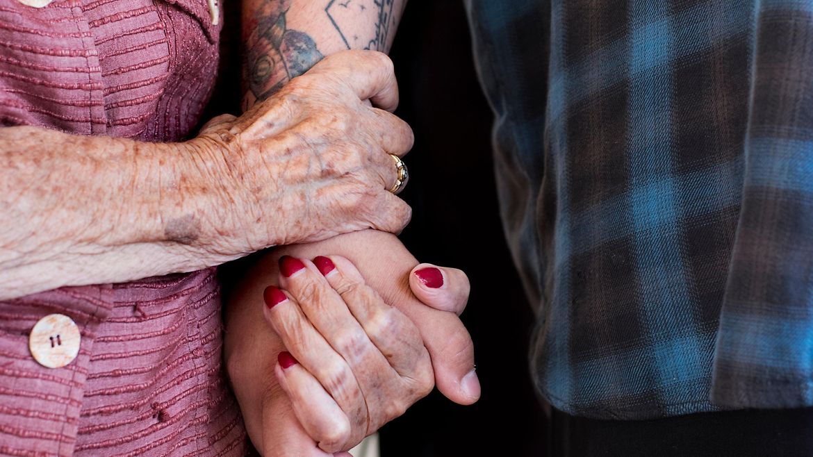 Eine ältere Frau hält sich am tätowierten Arm ihres Pflegers fest.