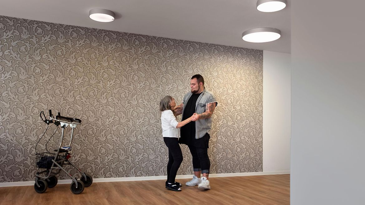 Eine kleine ältere Frau tanzt mit einem großen jungen Mann in einem Zimmer. An der Wand steht ein Rollator.