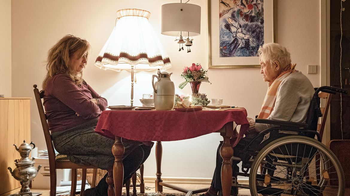 Eine ältere Frau im Rollstuhl sitzte mit einer jüngeren Frau an einem gedeckten Tisch.