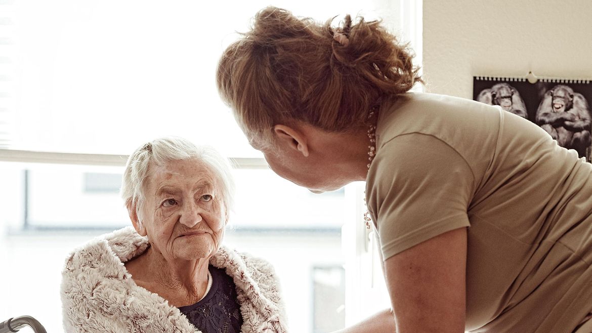Eine ältere Frau sitzt im Rollstuhl vor einem Pflegebett und schaut einer jüngeren Frau in die Augen.