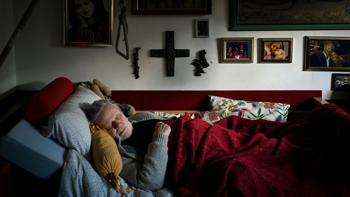 Ein Mann schläft in einem Bett vor einer Wand mit vielen Bilderrahmen.