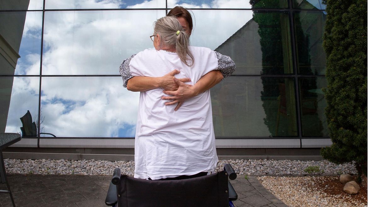Eine ältere Frau und eine Pflegerin umarmen sich vor einem Rollstuhl.