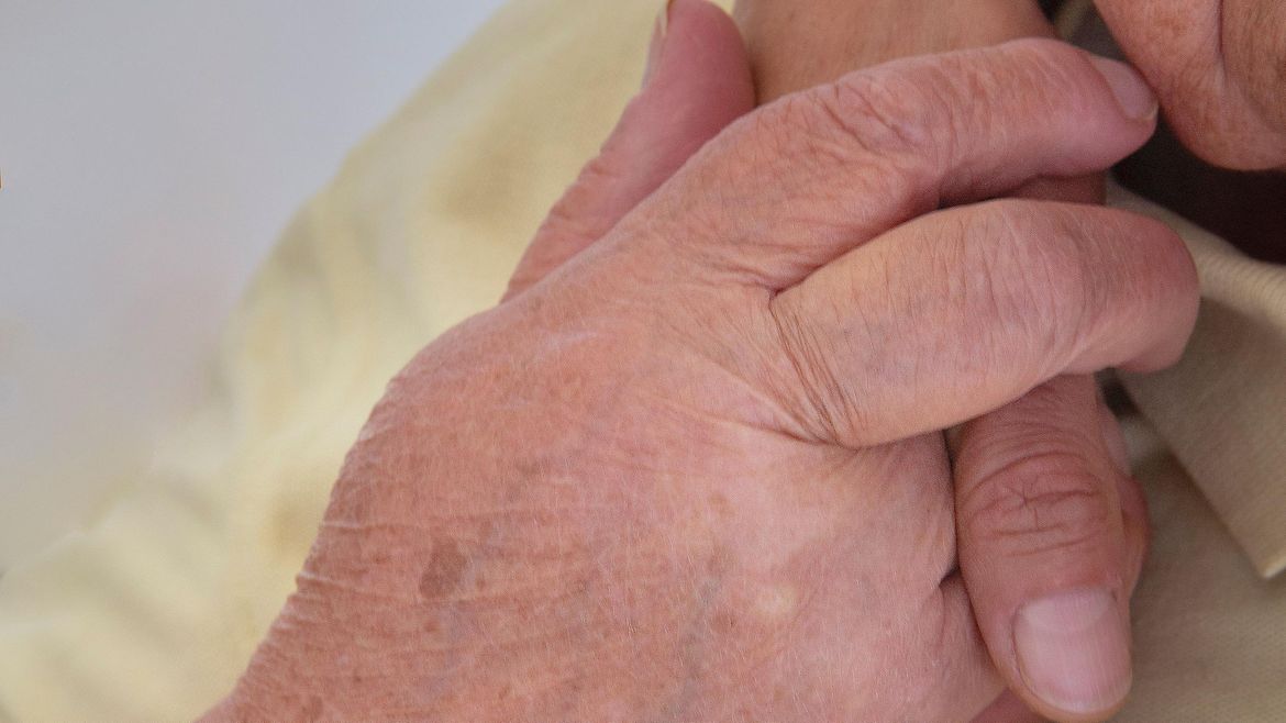 Eine ältere Person hält die Hand einer jüngeren, die um ihre Schulter liegt.