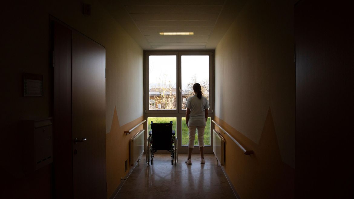 Eine Pflegerin steht neben einem leeren Rollstuhl vor einem Fenster.