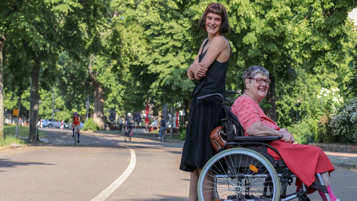 Eine junge Frau und eine ältere Frau im Rollstuhl stehen Rücken an Rücken auf einer Straße.