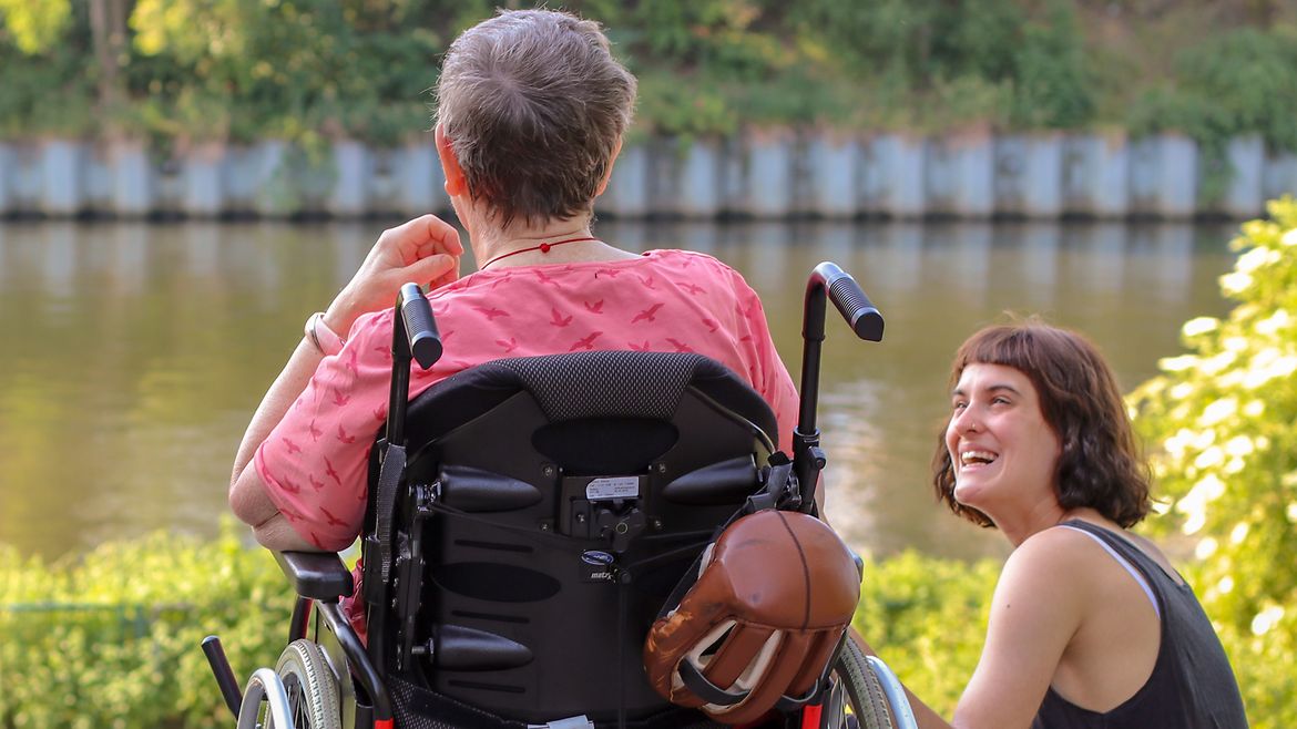 Eine alte Frau im Rollstuhl und eine junge Frau sitzen am Ufer eines Flusses.