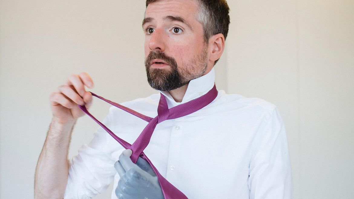 Ein bärtiger Mann mit einer Prothese am linken Arm bindet sich eine Krawatte.