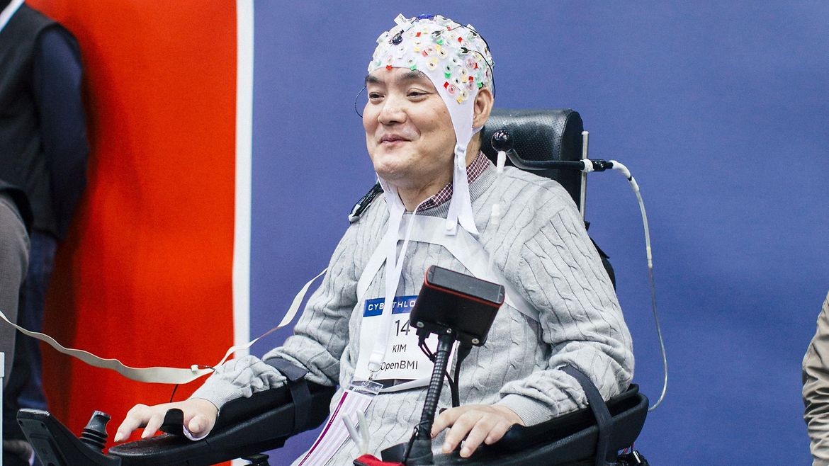 Ein asiatischer Mann im Rollstuhl vor einer Zuschauertribüne. Er trägt eine weiße Kappe an der verschiedenfarbige Elektroden befestigt sind.