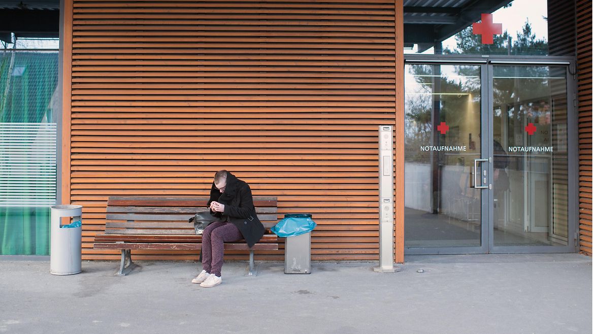 Eine junge Frau sitzt auf einer Bank vor einer Klinik.