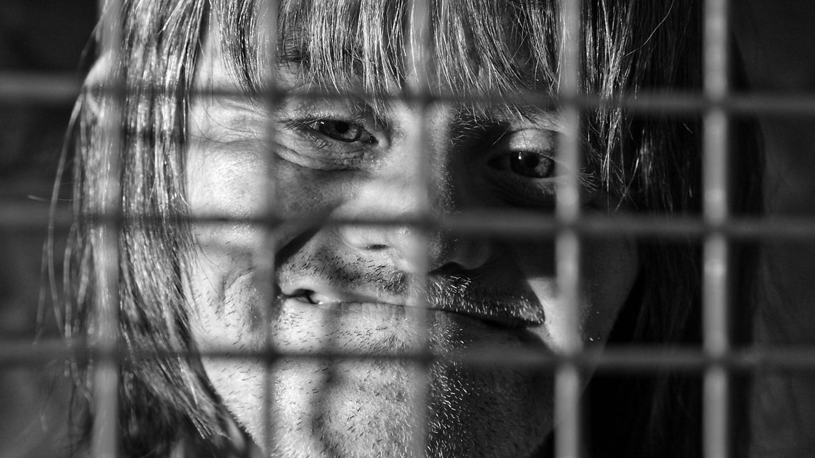 Ein Mann - mit phänotypischen Zügen der Trisomie 21 - lächelt, wobei er hinter einem Gitter zu sehen ist. 