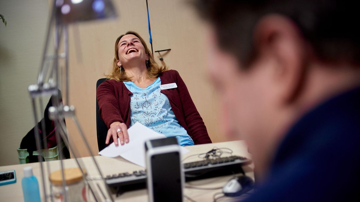 Eine Frau sitzt am Schreibtisch einem Mann gegenüber und lacht.