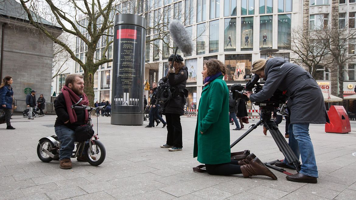 Ein kleinwüchsiger Mann sitzt auf einem Roller und wird von einem Fernsehteam in einer Einkaufsstraße gefilmt.