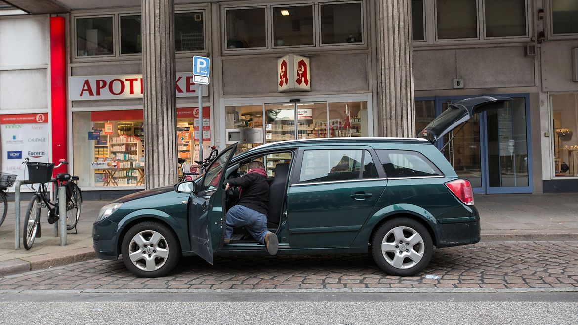 Ein kleinwüchsiger Mann steigt in ein Auto, das vor einer Apotheke steht.