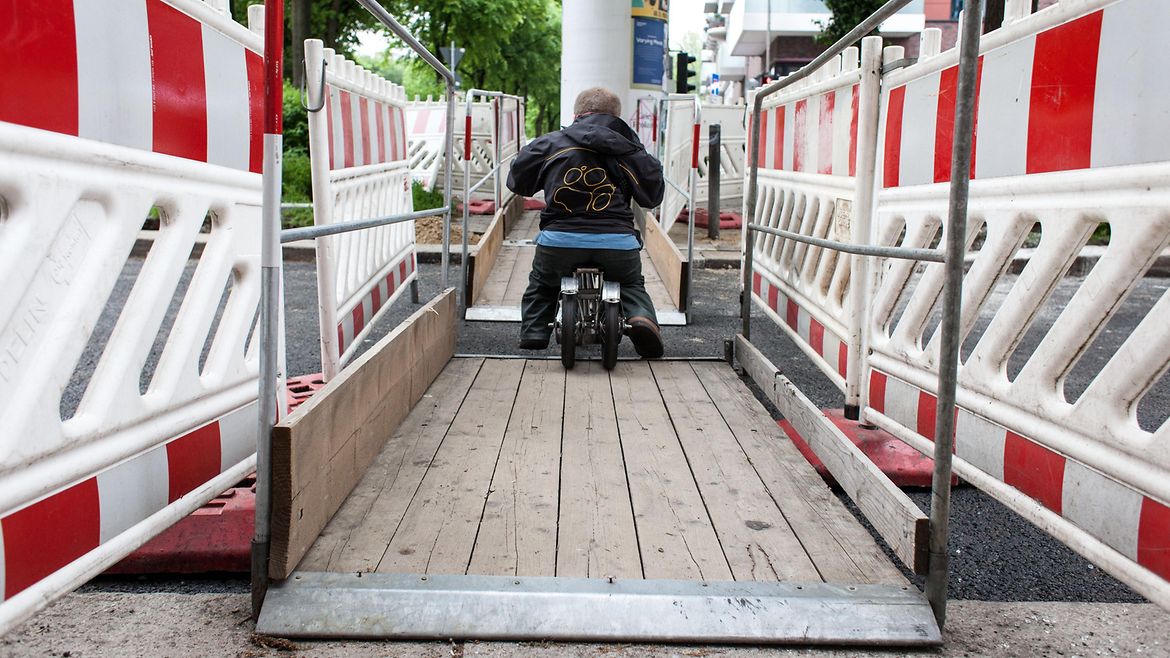 Ein kleinwüchsiger Mann fährt auf einem Roller über eine Baustelle auf einer Straße.