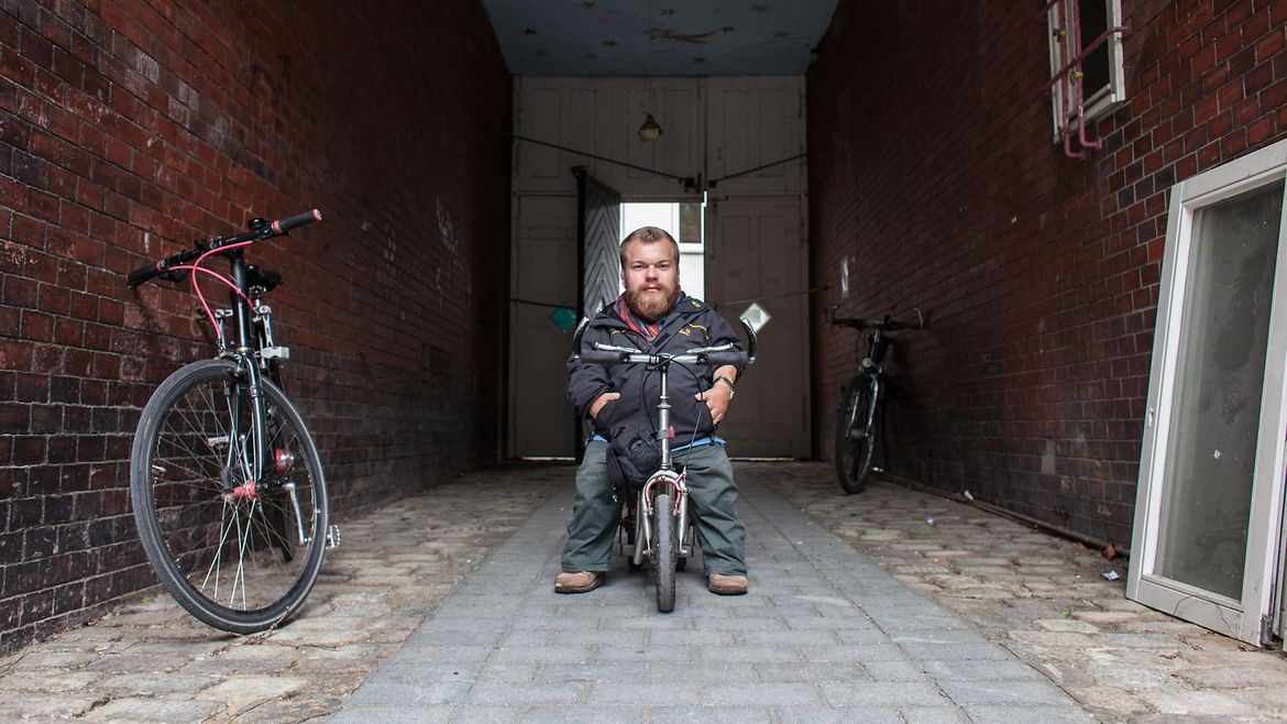 Ein kleinwüchsiger Mann sitzt auf einem Roller in einem Hauseingang.